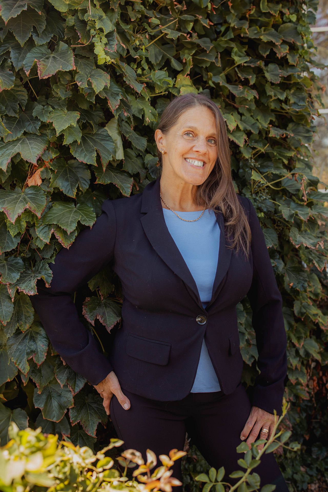 Carrie DeGrandmont, in a navy blazer and light blue top, smiling in front of a green ivy wall